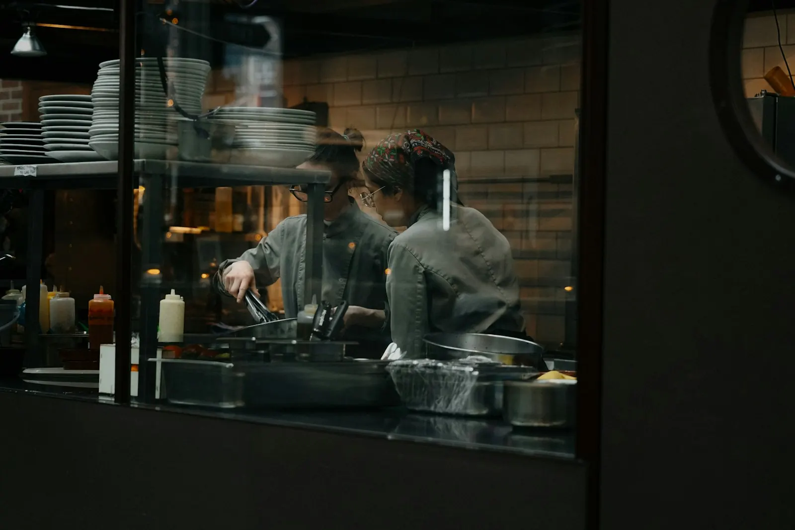 Twee koks aan het werk in een open keuken, gezien door het raam van de pass